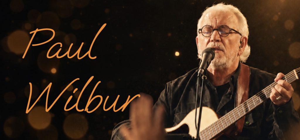 Paul Wilbur performing with an acoustic guitar on stage, orange cursive name text on a dark bokeh background.